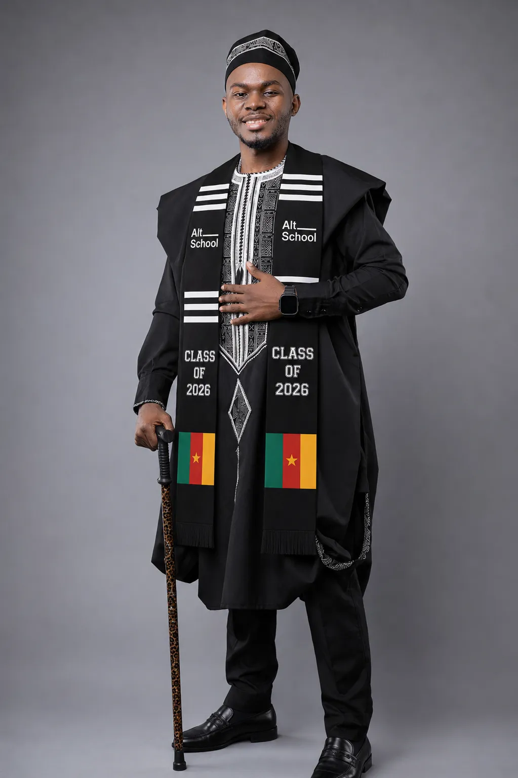Michel Tegofack en tenue traditionnelle noire et coiffe, portant l'étole AltSchool « Class of 2026 » aux drapeaux du Cameroun, une canne à la main, sur fond gris.